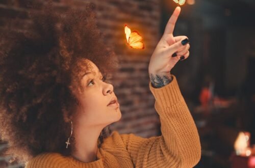 A woman with afro hair in a cozy setting playfully interacts with butterflies indoors.