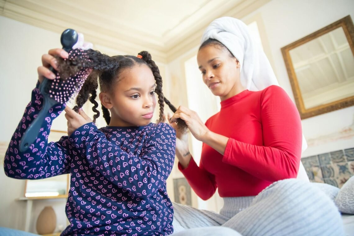 A mother braids her daughter's hair at home, emphasizing bonding and ethnic hair care.