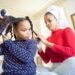 A mother braids her daughter's hair at home, emphasizing bonding and ethnic hair care.